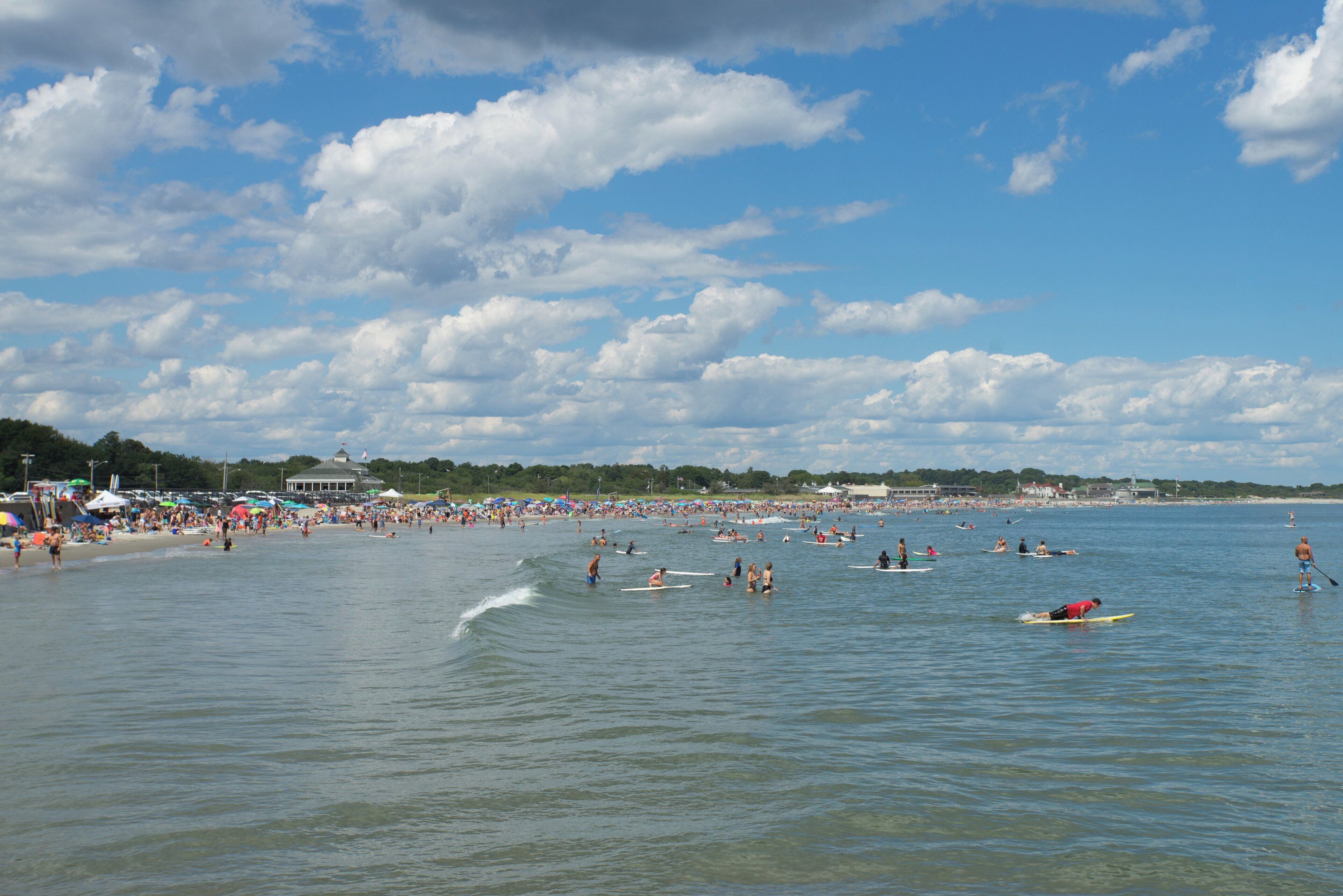 A beach with people engaging in water activities, a clear sky, and a distant shoreline with buildings.
