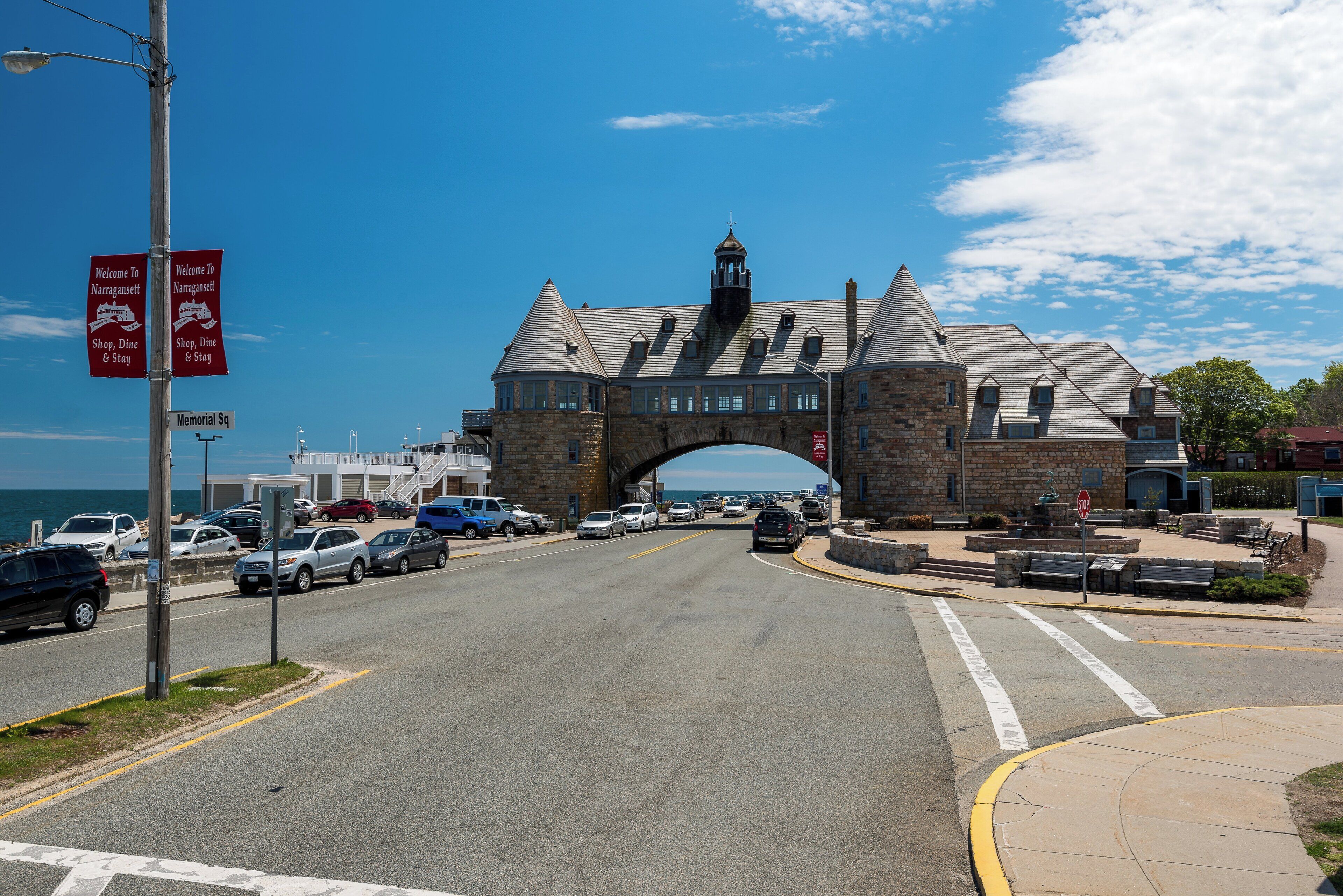 A historic stone building with a large arched entrance, a clock tower, and a sign welcoming visitors to Narragansett.