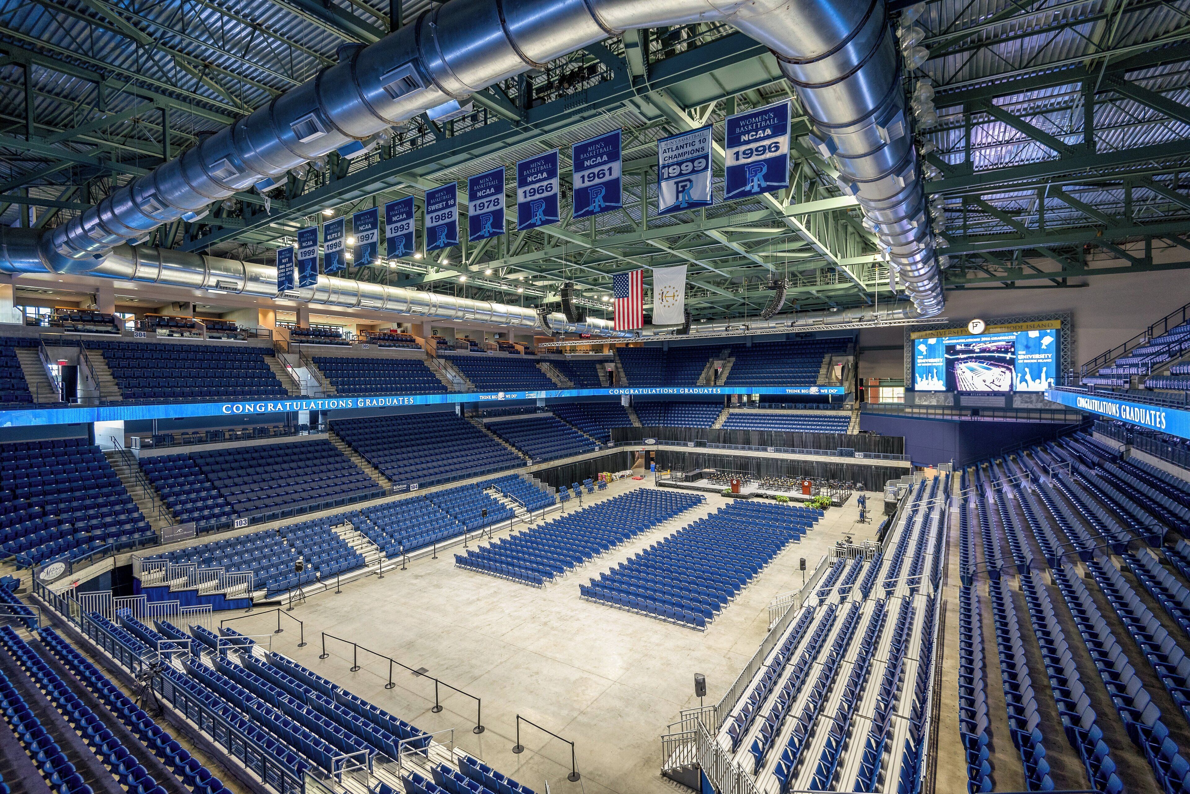 Un vaste stade intérieur doté de plusieurs niveaux de gradins, d’une scène centrale et d’un plafond élevé laissant apparaître des poutres de soutien et des conduits de ventilation.