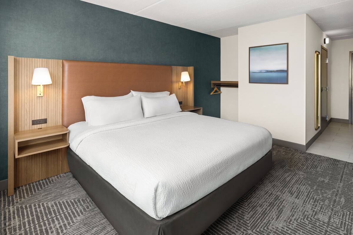 A hotel room with a large bed, two bedside tables with lamps, a wooden headboard, a wall-mounted picture, and a carpeted floor.