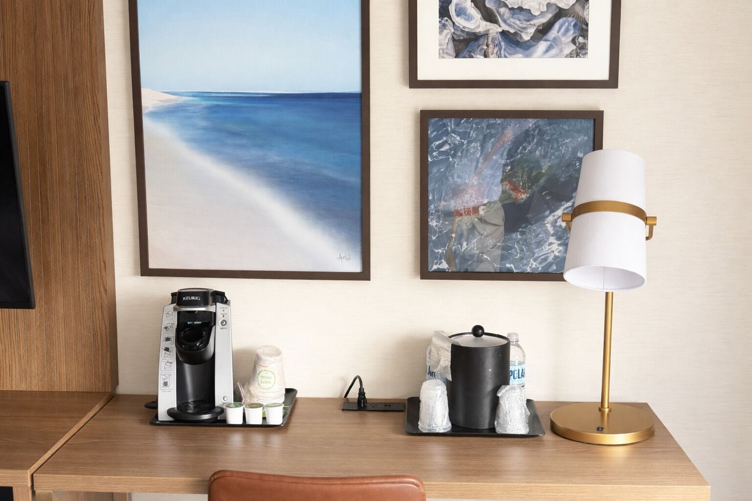 A hotel room with a wooden desk, a coffee machine, a lamp, and framed artwork on the wall.