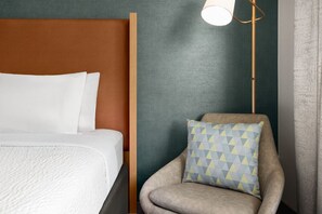 Un lit avec une literie blanche, une tête de lit marron, un fauteuil gris avec un coussin à motifs et une lampe sur pied.