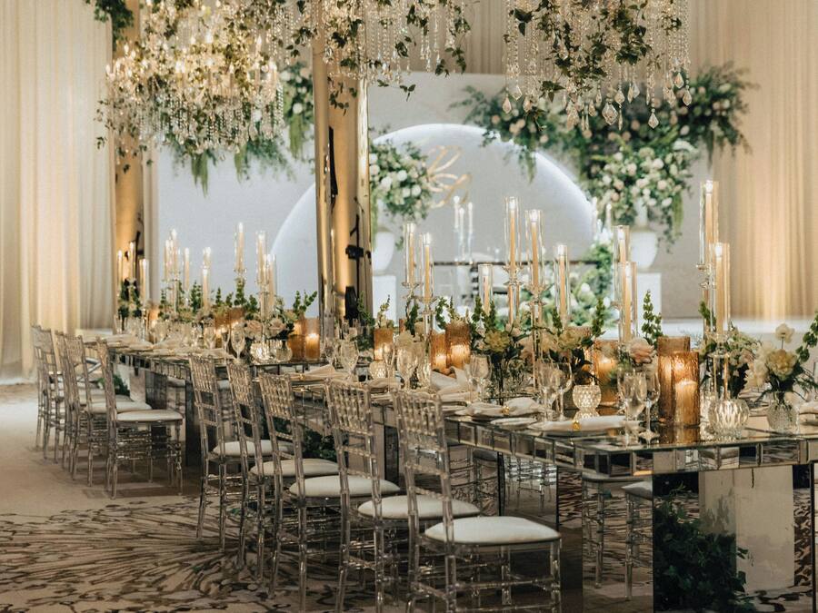 A banquet hall with a long table set for a formal event, surrounded by chairs and adorned with floral arrangements and candles.
