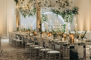 A banquet hall with a long table set for a formal event, surrounded by chairs and adorned with floral arrangements and candles.