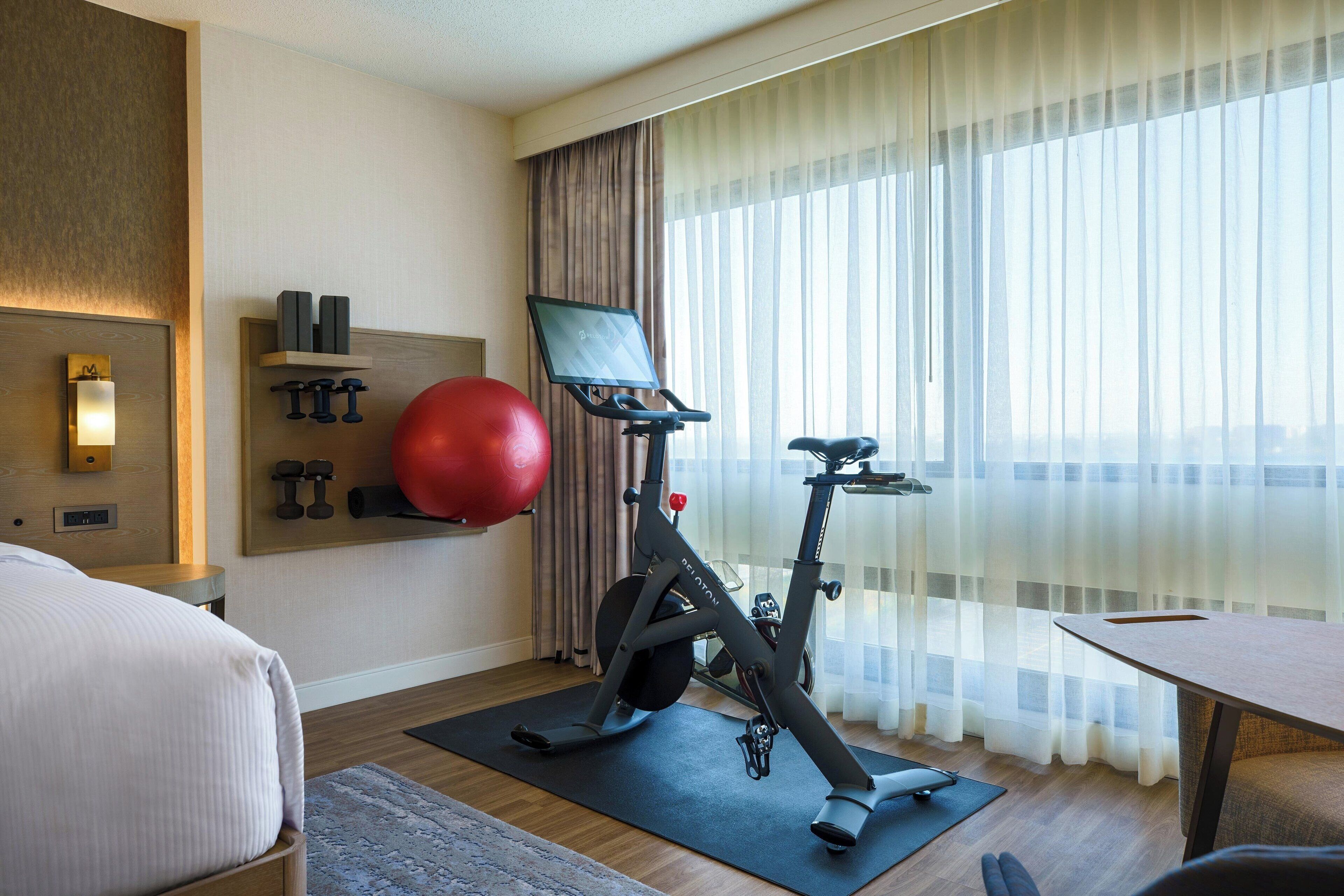 A hotel room with a bed, exercise equipment, a desk, and a large window with curtains.