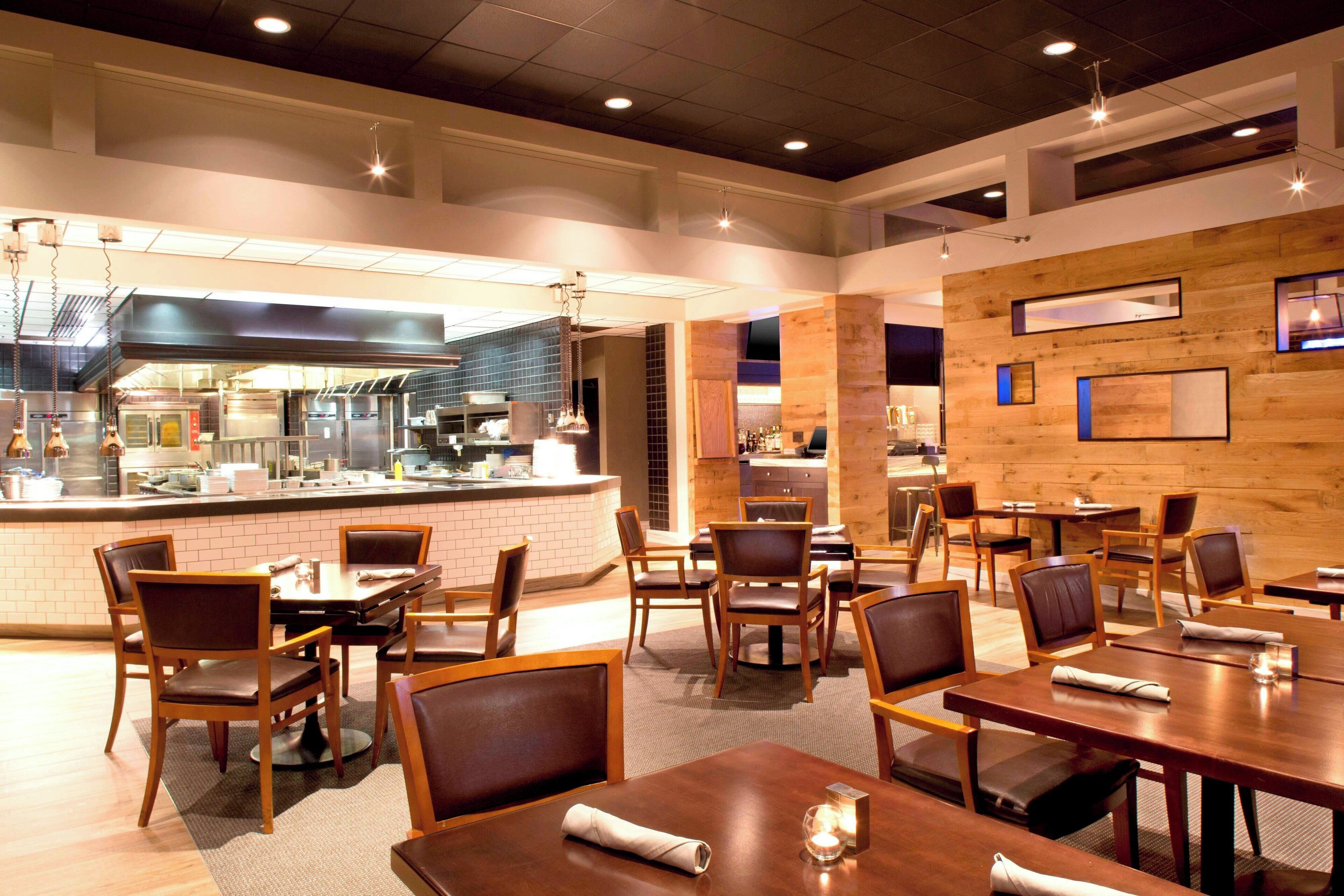 A modern restaurant interior with wooden tables and chairs, a kitchen area, and a bar.