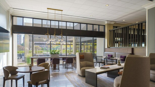 Eine großzügige Lobby mit modernem Design, ausgestattet mit einer geräumigen Sitzecke, Sofas und Sesseln, einem Couchtisch sowie einem Esstisch mit Stühlen. Große Fenster bieten einen Blick ins Freie.