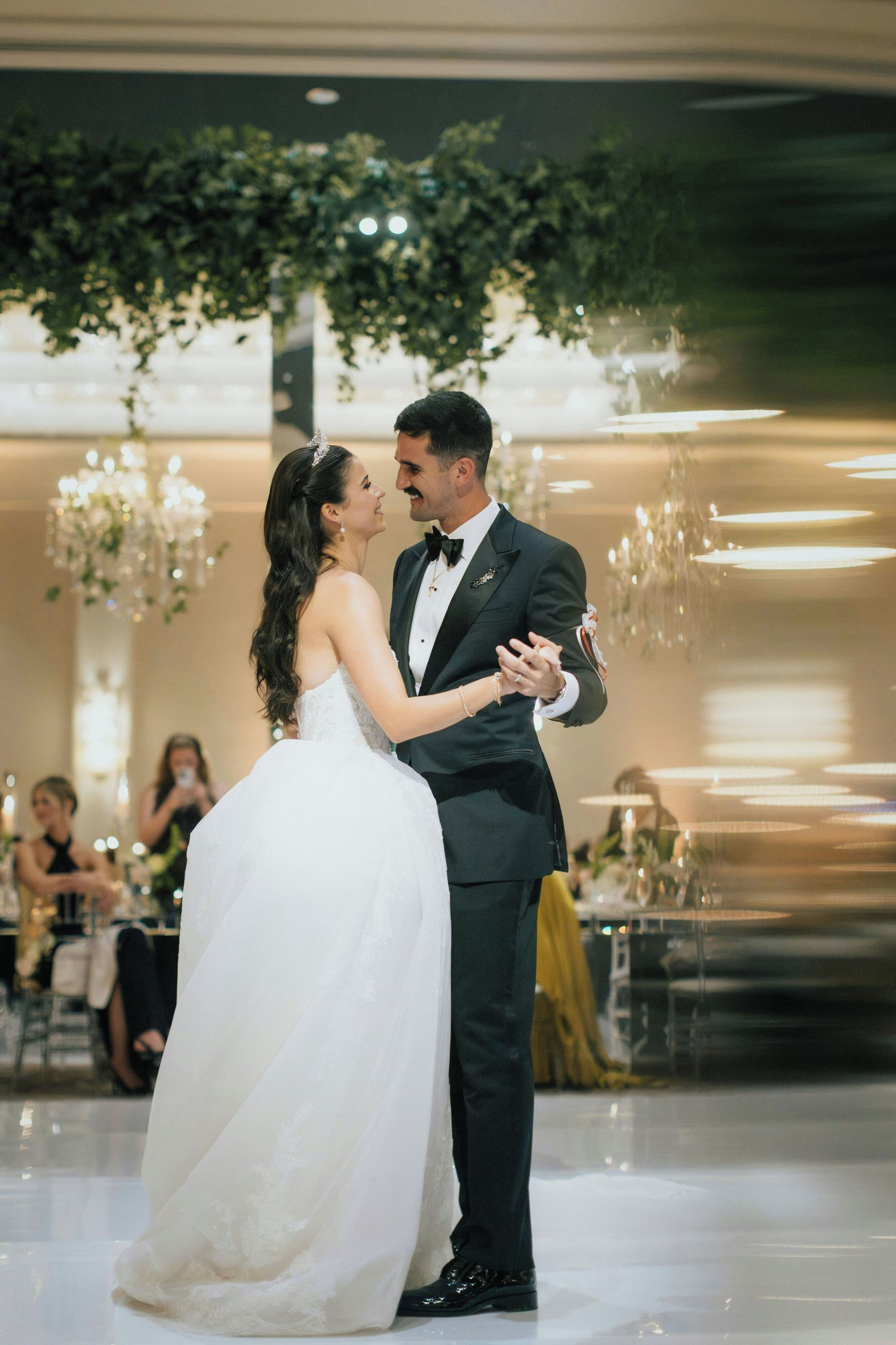 A couple dancing in a formal event setting with a chandelier and floral decorations.