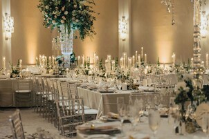 A banquet hall with long tables set for a formal event, featuring clear chairs, white tablecloths, and floral centerpieces.