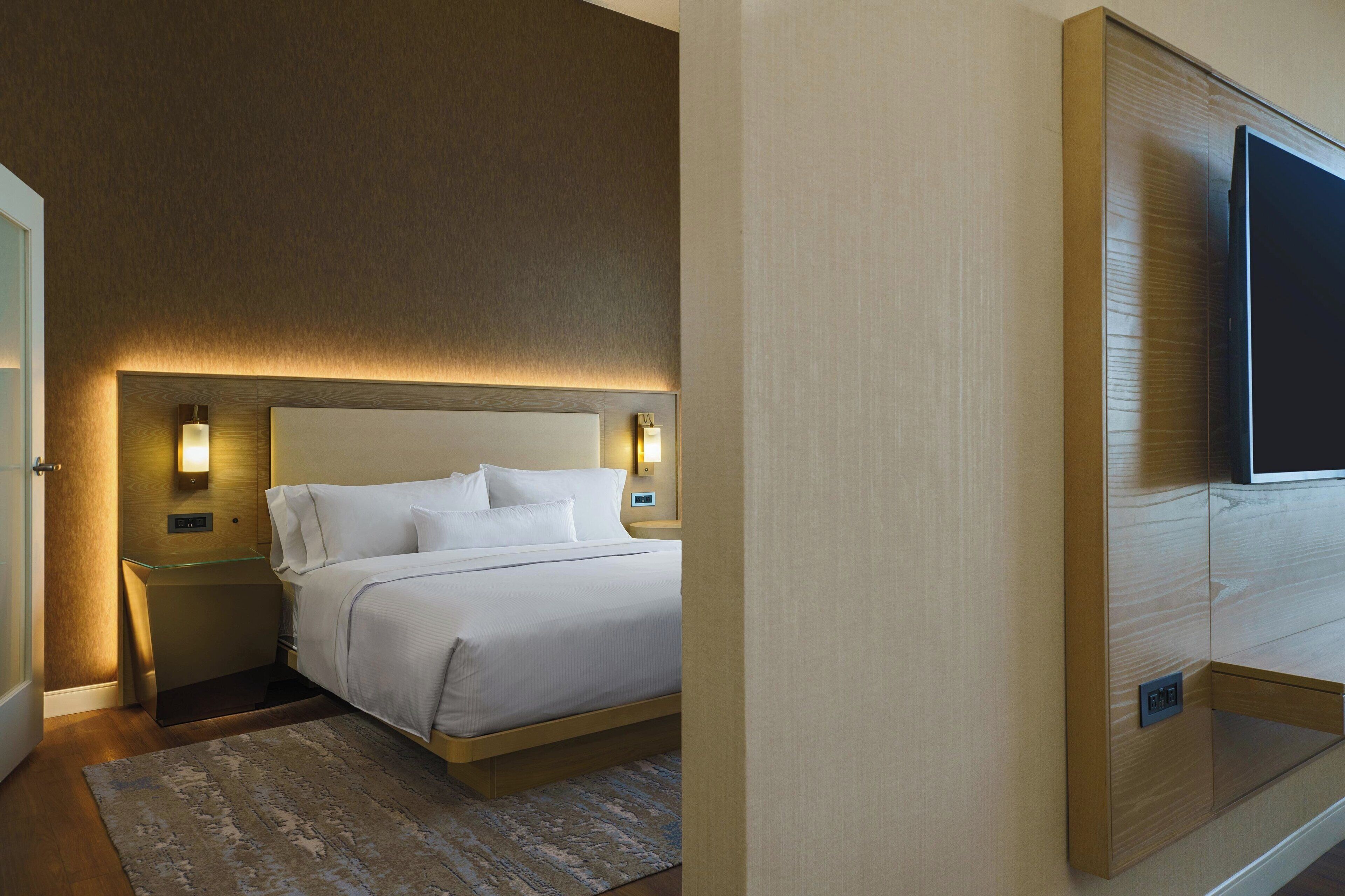 Habitación de hotel con cama, lámparas de noche, televisor y cabecera de madera.