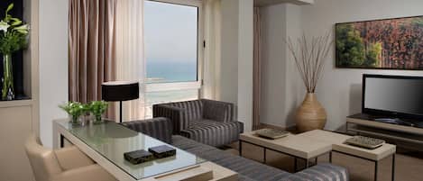 A modern hotel room with a glass coffee table, two sofas, a TV, and a large window with a view of the sea.