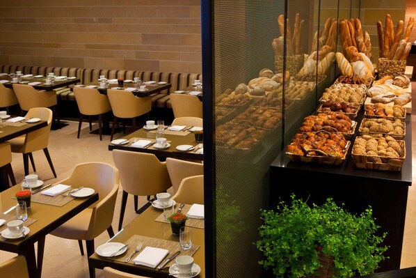 A dining area with tables set for service, a variety of breads and pastries displayed, and a seating area with cushioned chairs.