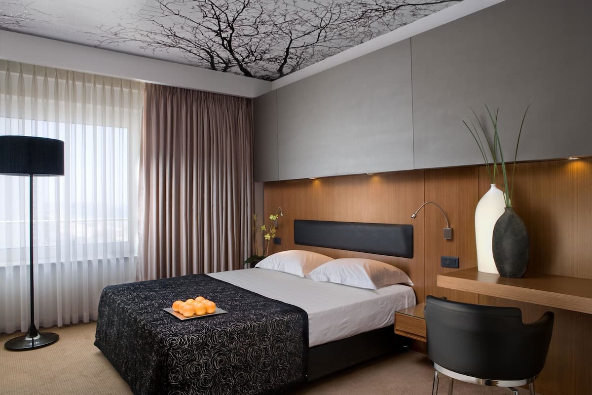 A modern hotel room with a bed, a desk, a chair, a lamp, and a vase with plants.