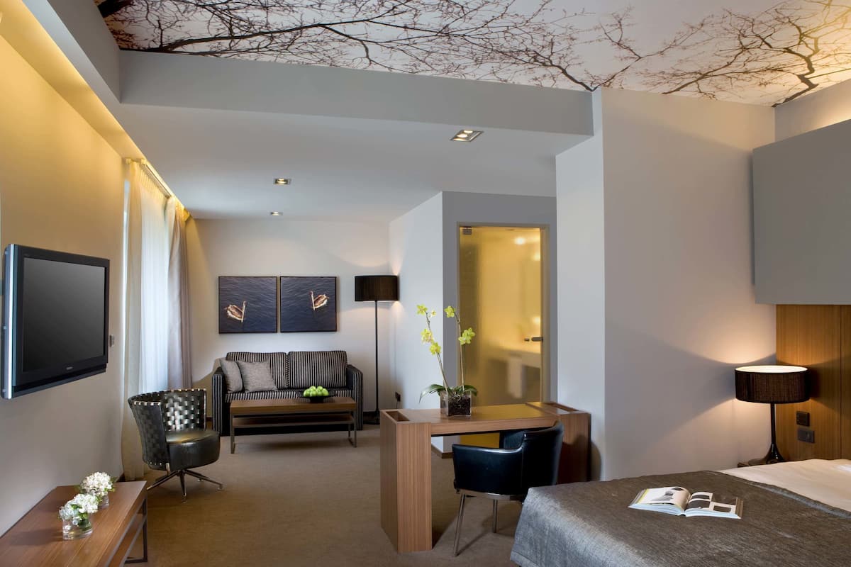 A modern hotel room with a flat-screen TV, a sofa, a desk, a chair, a bed, and a nightstand.