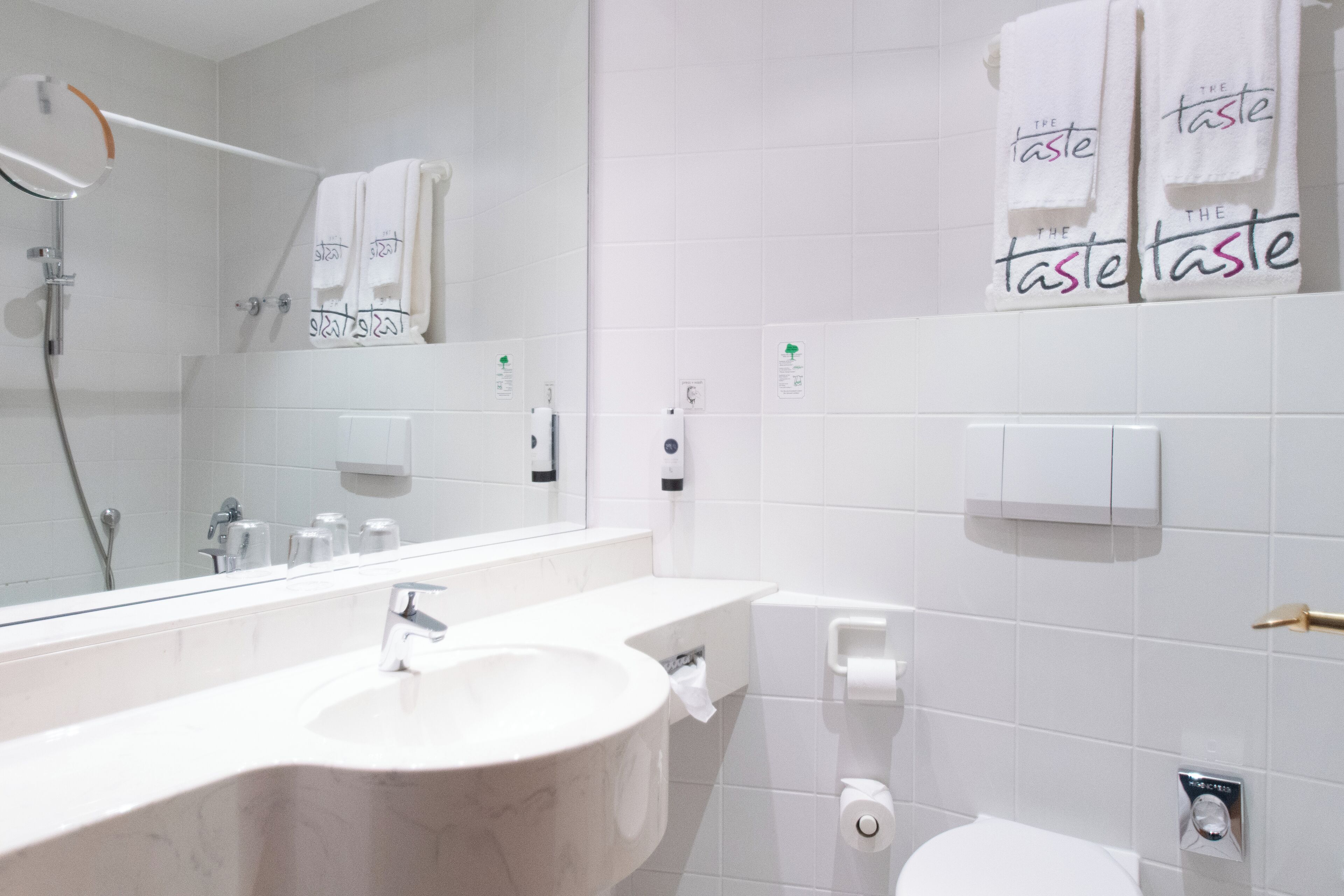 A modern bathroom with a large mirror, a sink, and a toilet. There are towels hanging on the wall and a showerhead visible.