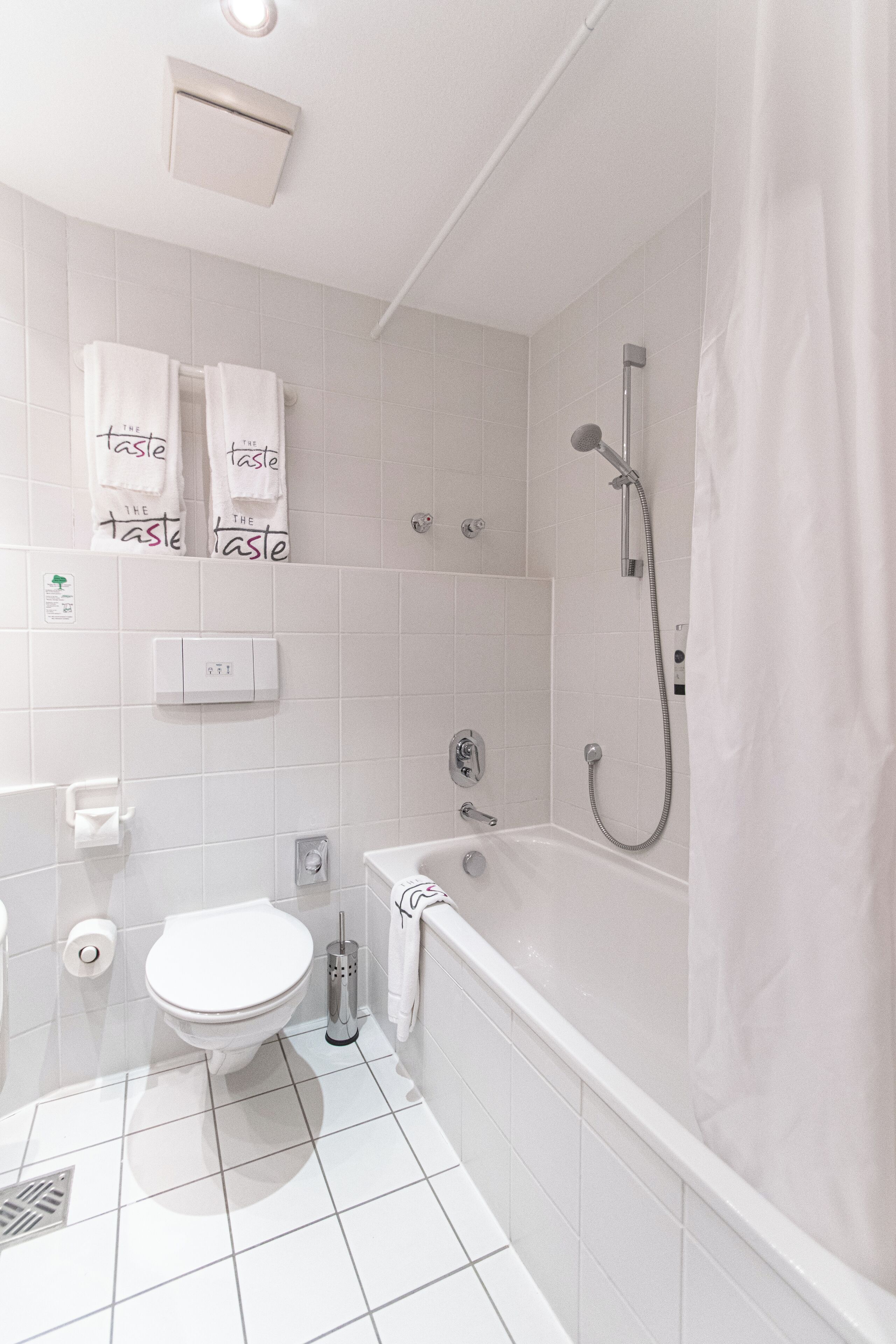 Un baño moderno con inodoro blanco, bañera y ducha. Toallas colgadas en la pared y un dispensador de papel higiénico.