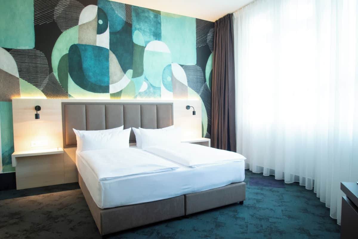 A modern hotel room with a large bed, a headboard with integrated lighting, a wall art piece, and a view of the outdoors through sheer curtains.