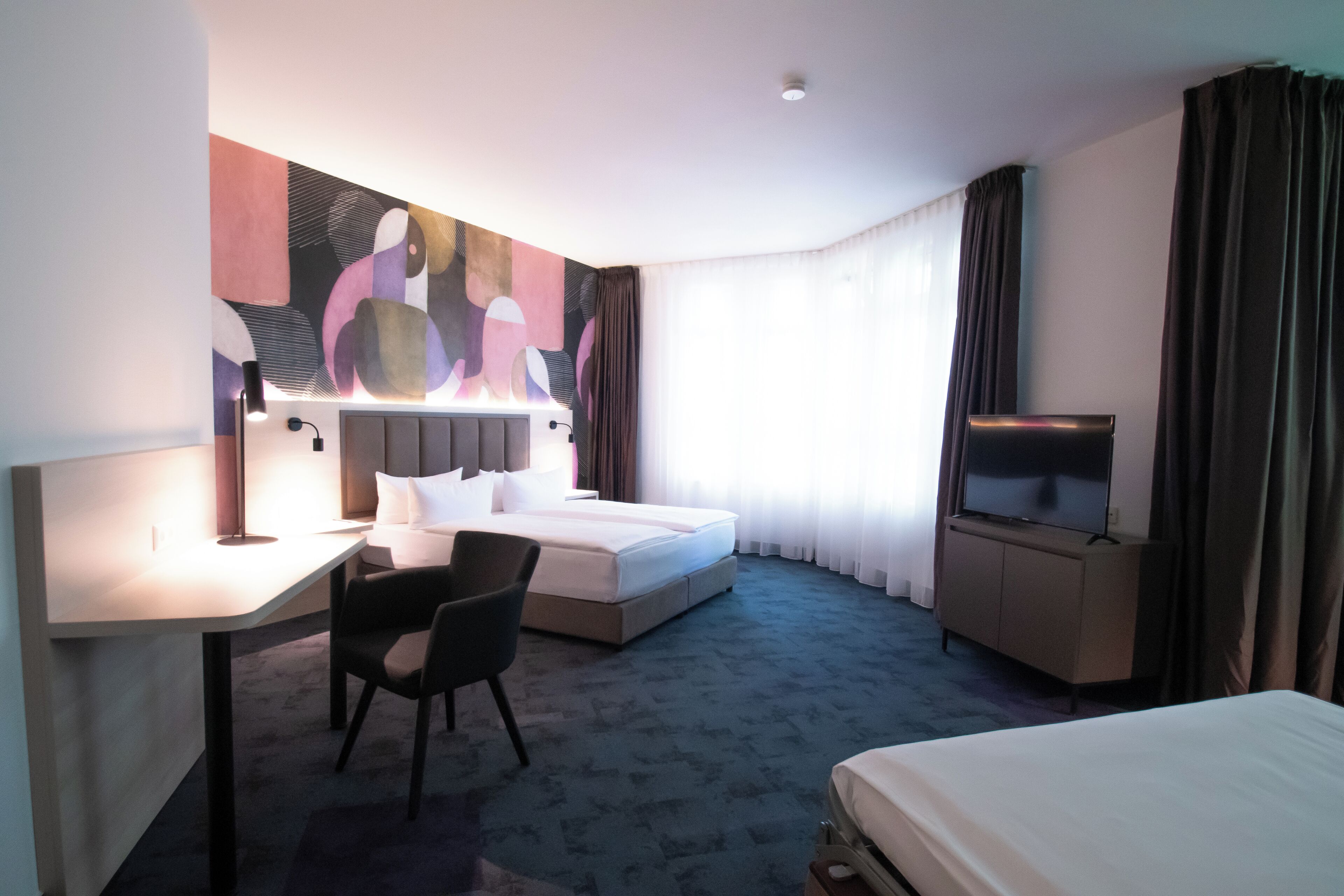 Habitación de hotel con cama, escritorio, silla, televisor y un cuadro abstracto grande en la pared.