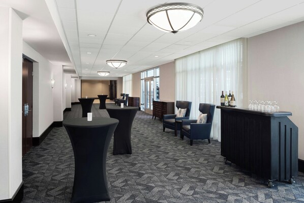 A hotel lobby with a long corridor, round tables, seating areas, and a bar with wine glasses.