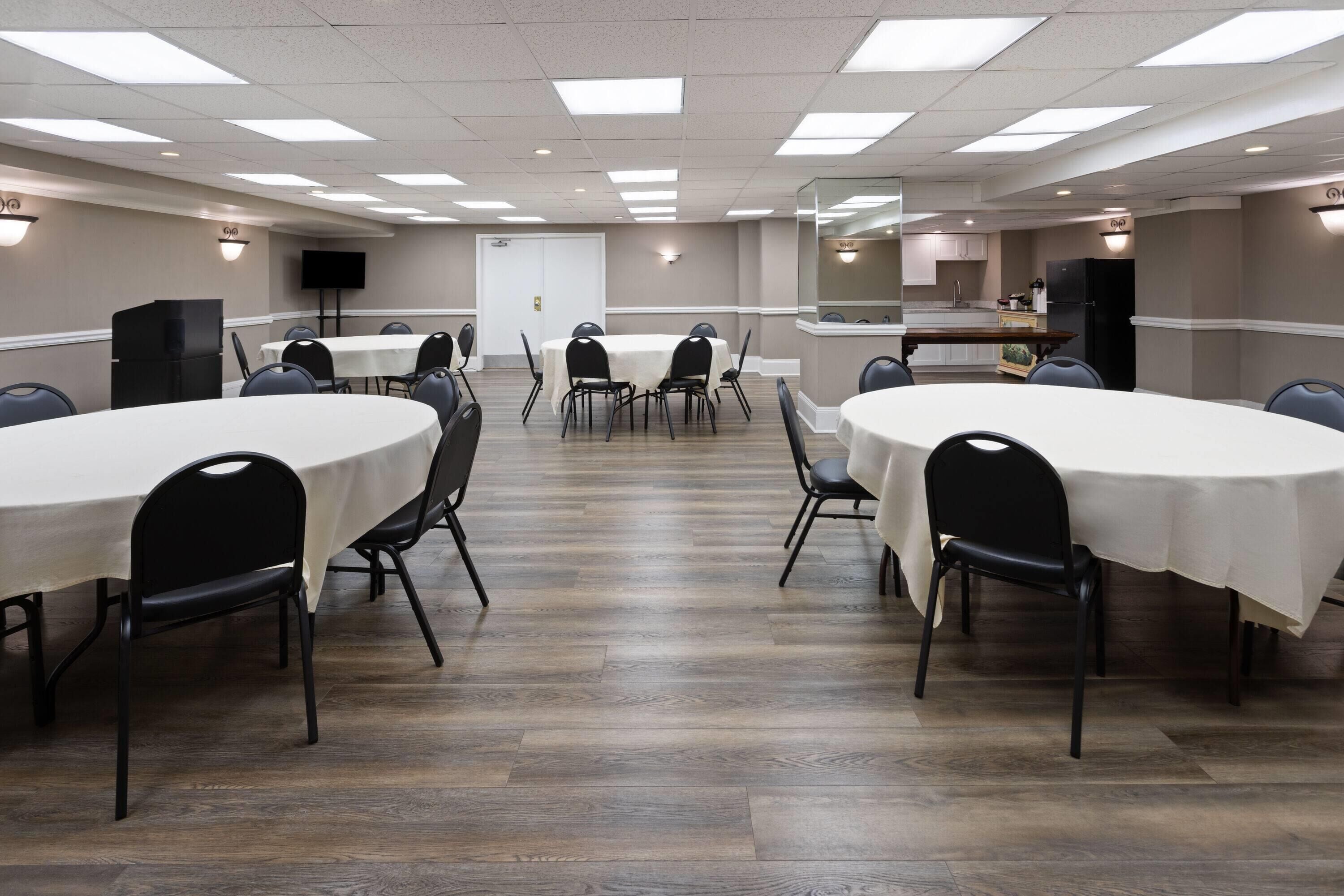 Une salle de conférence équipée de plusieurs tables et chaises, d’un coin cuisine et d’un téléviseur fixé au mur.
