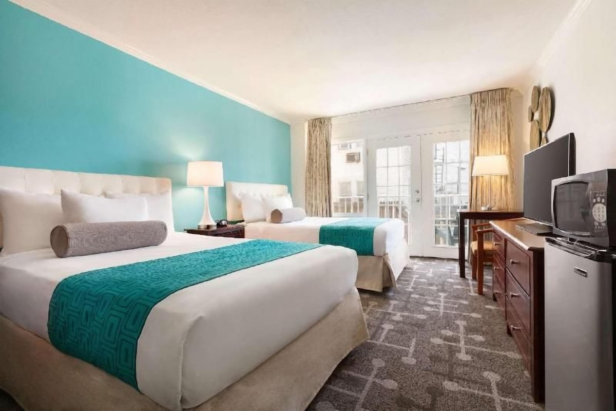 A hotel room with two beds, a desk, a television, and a view of the city.
