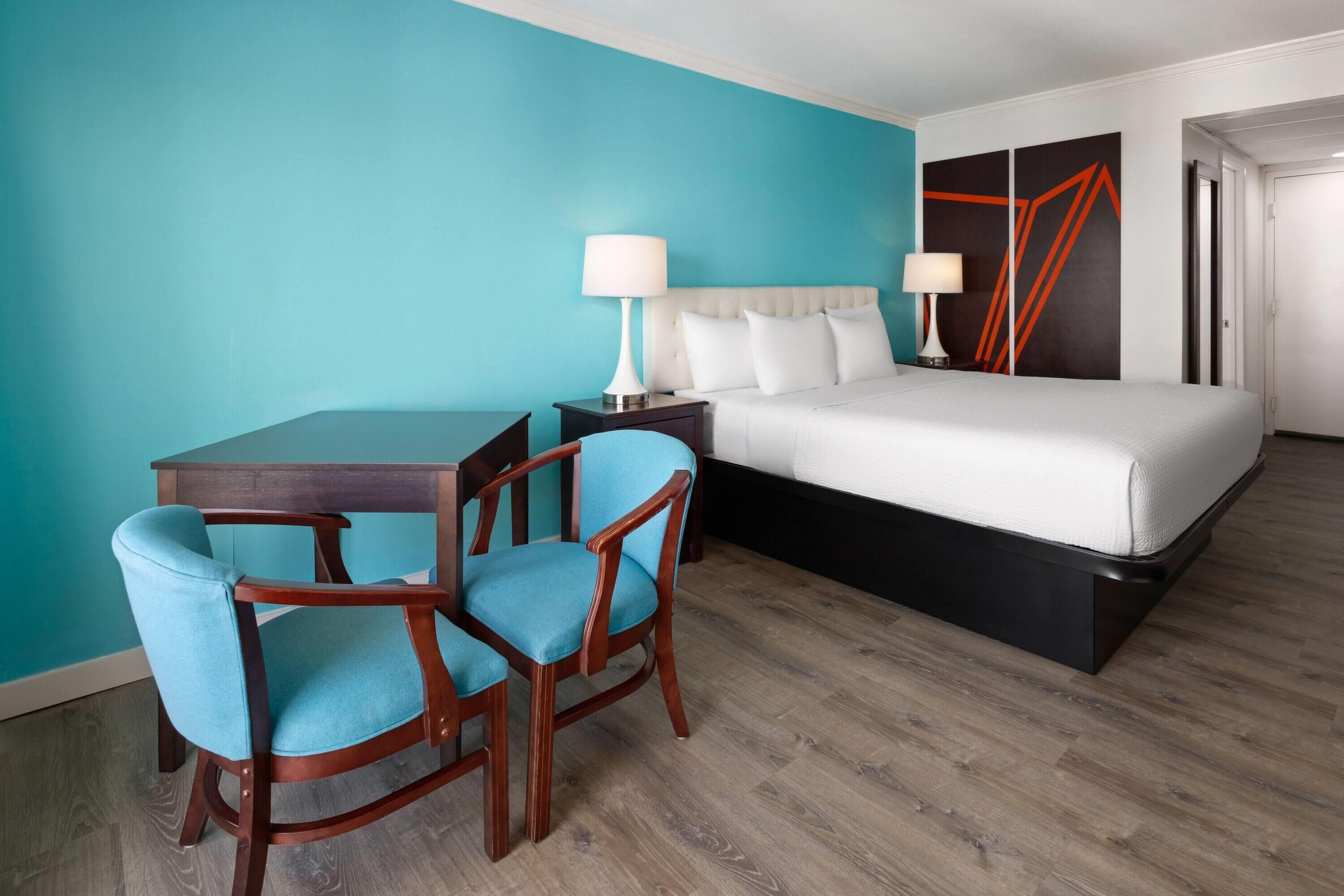 A hotel room with a bed, two bedside tables with lamps, a desk with two chairs, and a teal-colored wall.