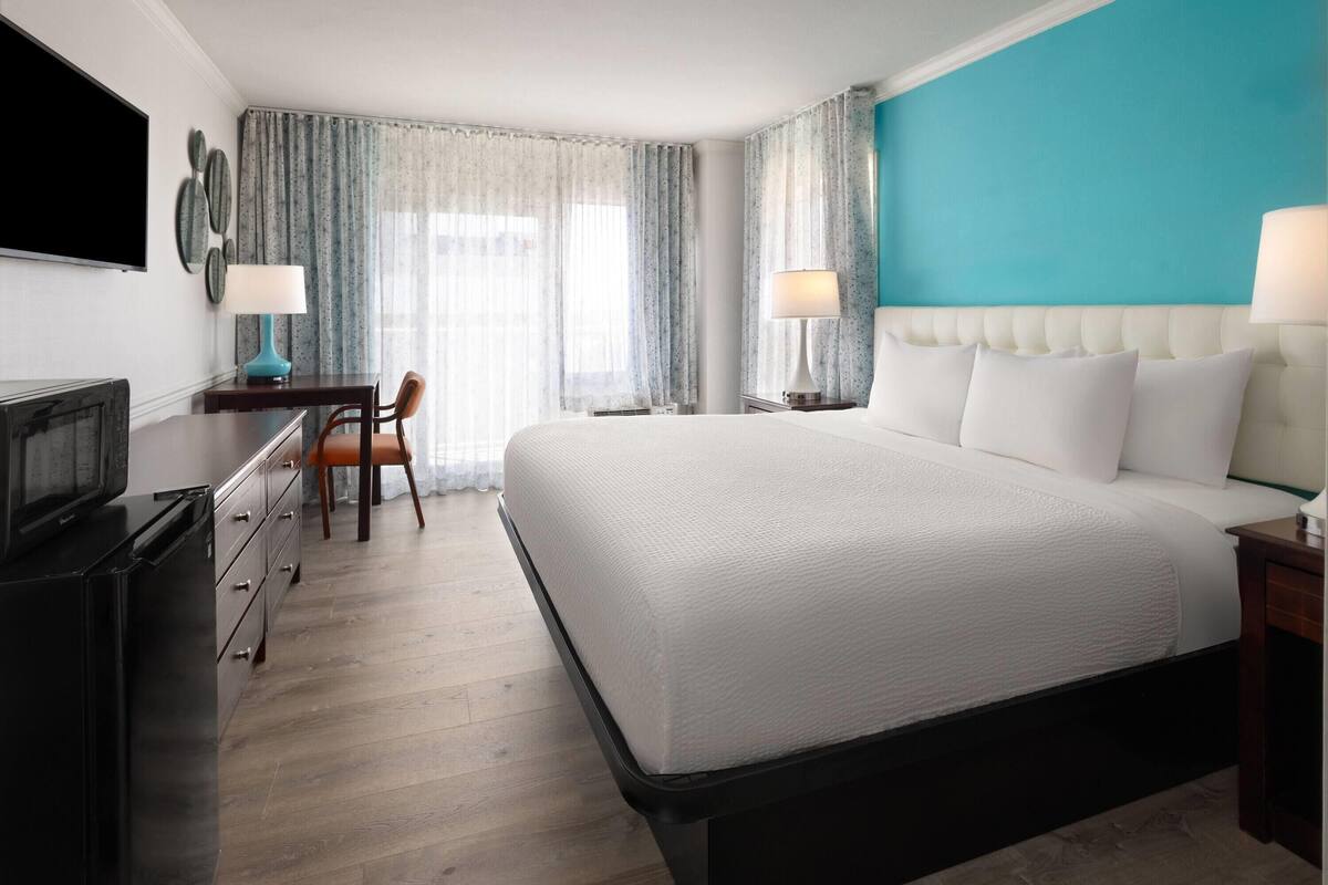 Une chambre d’hôtel avec un grand lit, un bureau avec une chaise, une télévision et une fenêtre avec des rideaux.