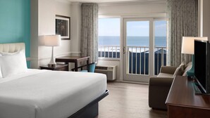 A hotel room with a bed, a desk, a chair, a TV, and a view of the ocean through a sliding door.