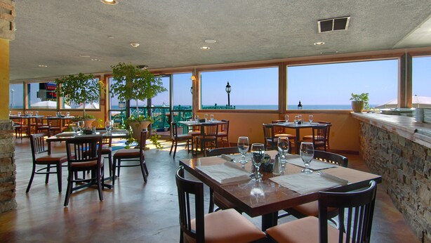 A restaurant with multiple tables and chairs, large windows offering a view of the sea, and a stone wall on one side.