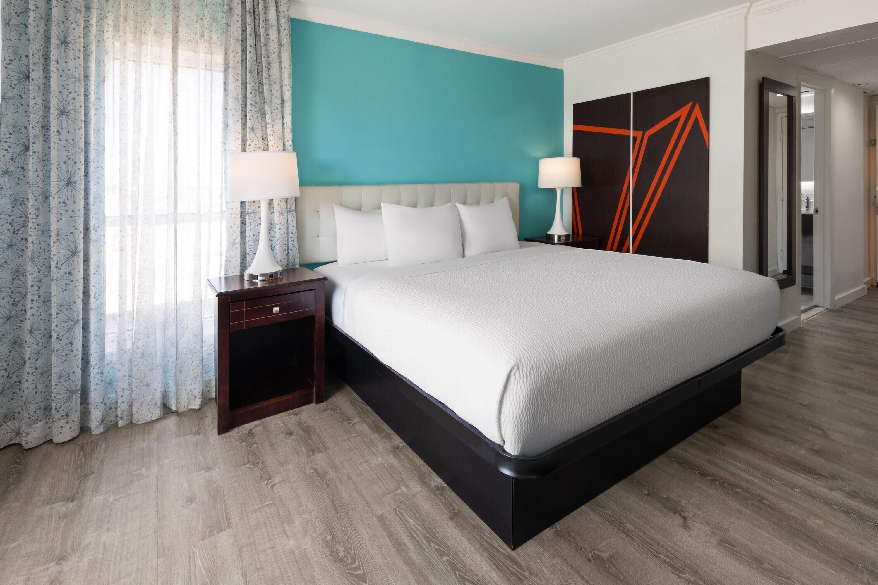 A modern hotel room with a large bed, two bedside tables with lamps, a teal accent wall, and a wooden floor.