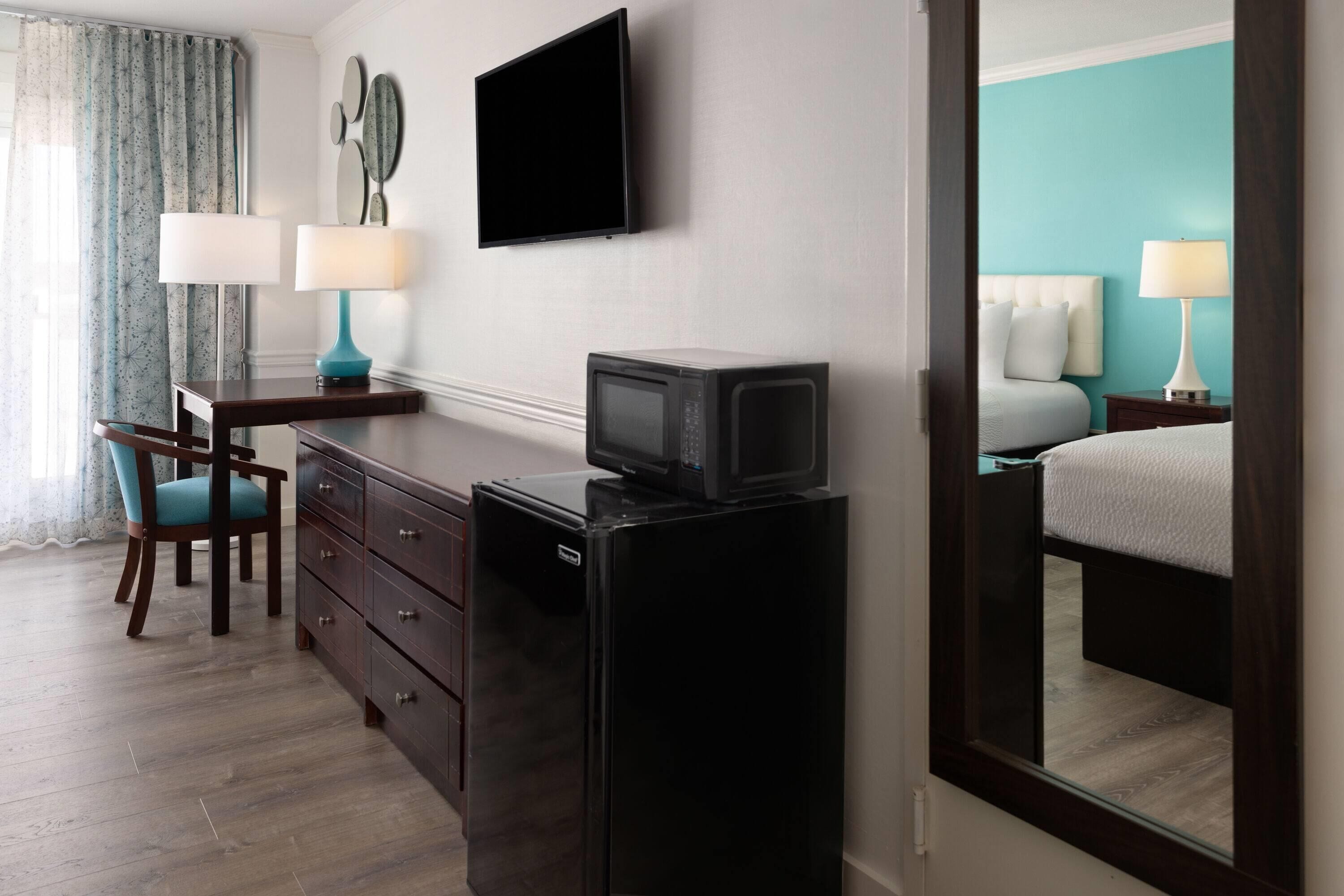 A hotel room with a bed, a desk with a chair, a television, a refrigerator, and a mirror.
