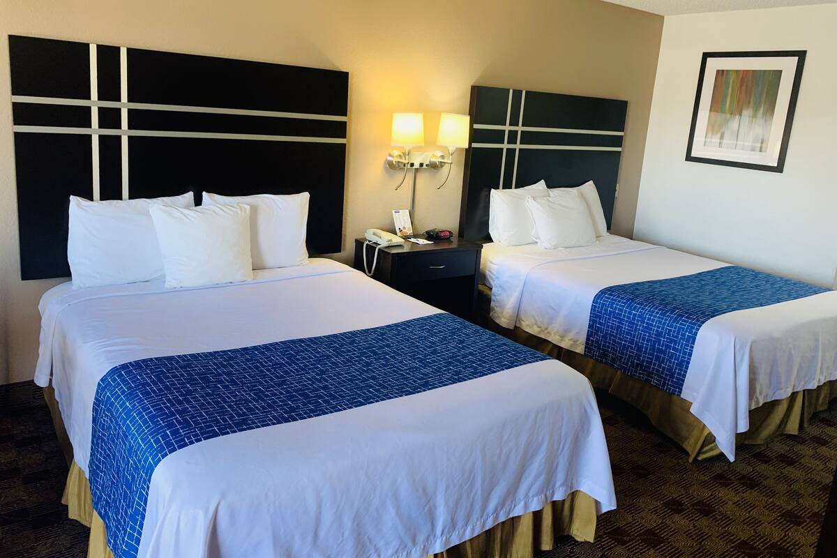A hotel room with two beds, each with white linens and blue bedspreads. There is a nightstand with a lamp and a phone between the beds. The room has a modern design with a large black headboard and a framed picture on the wall.