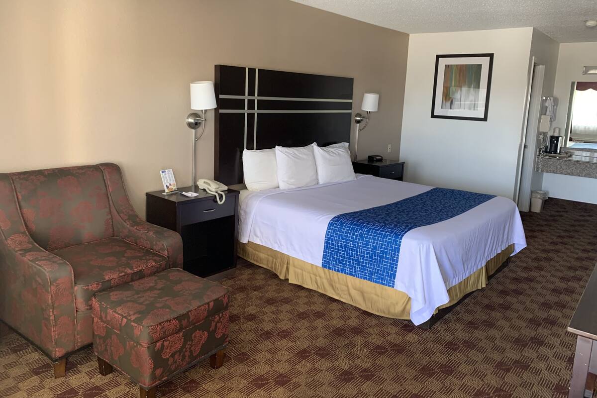 A hotel room with a bed, a nightstand, a lamp, a chair, and a bathroom visible through an open door.