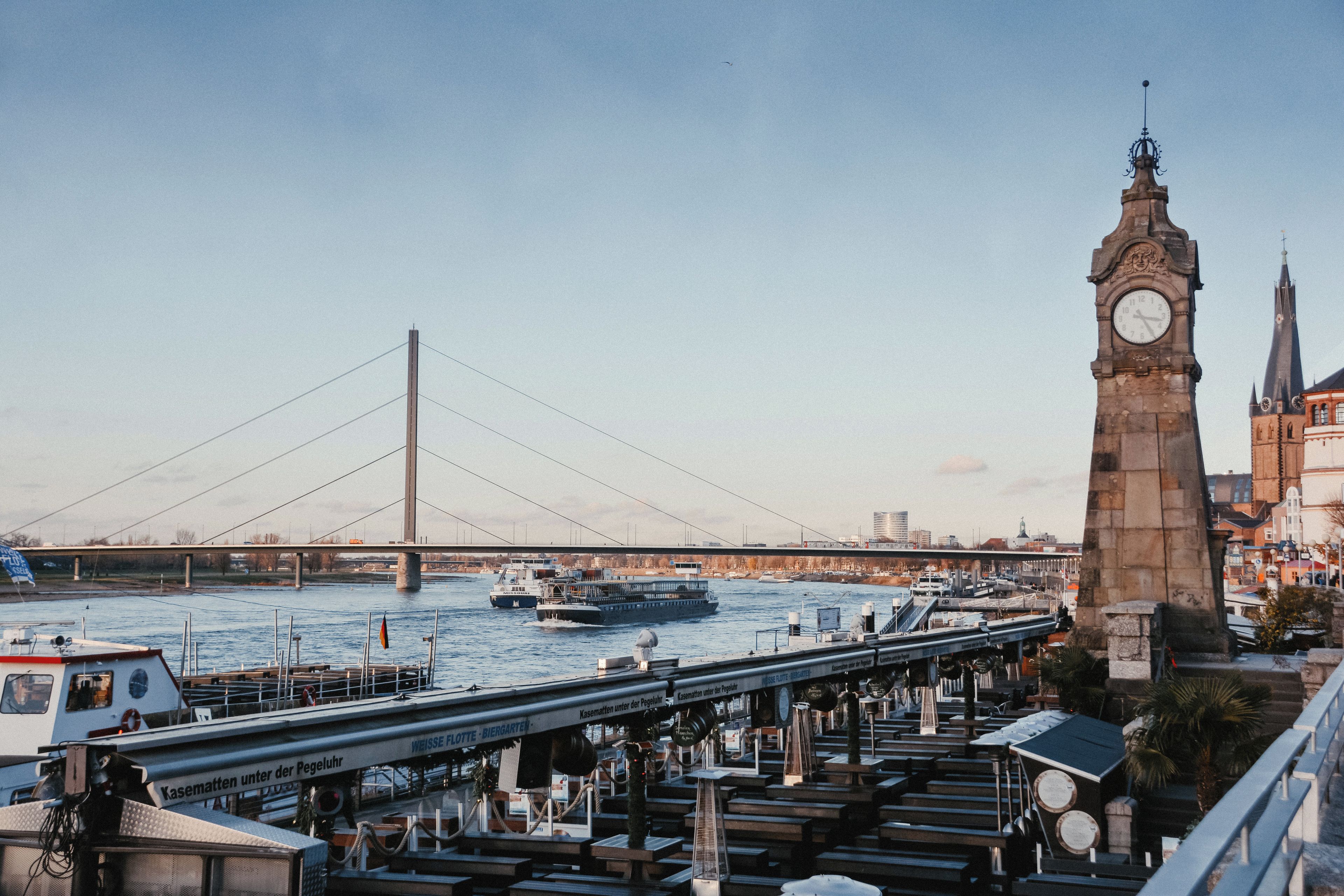 Vista junto al río con un puente, barcos y una torre del reloj.
