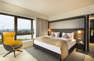 Una habitación de hotel moderna con una cama grande, un sillón amarillo y vistas a un paisaje urbano a través de amplios ventanales.