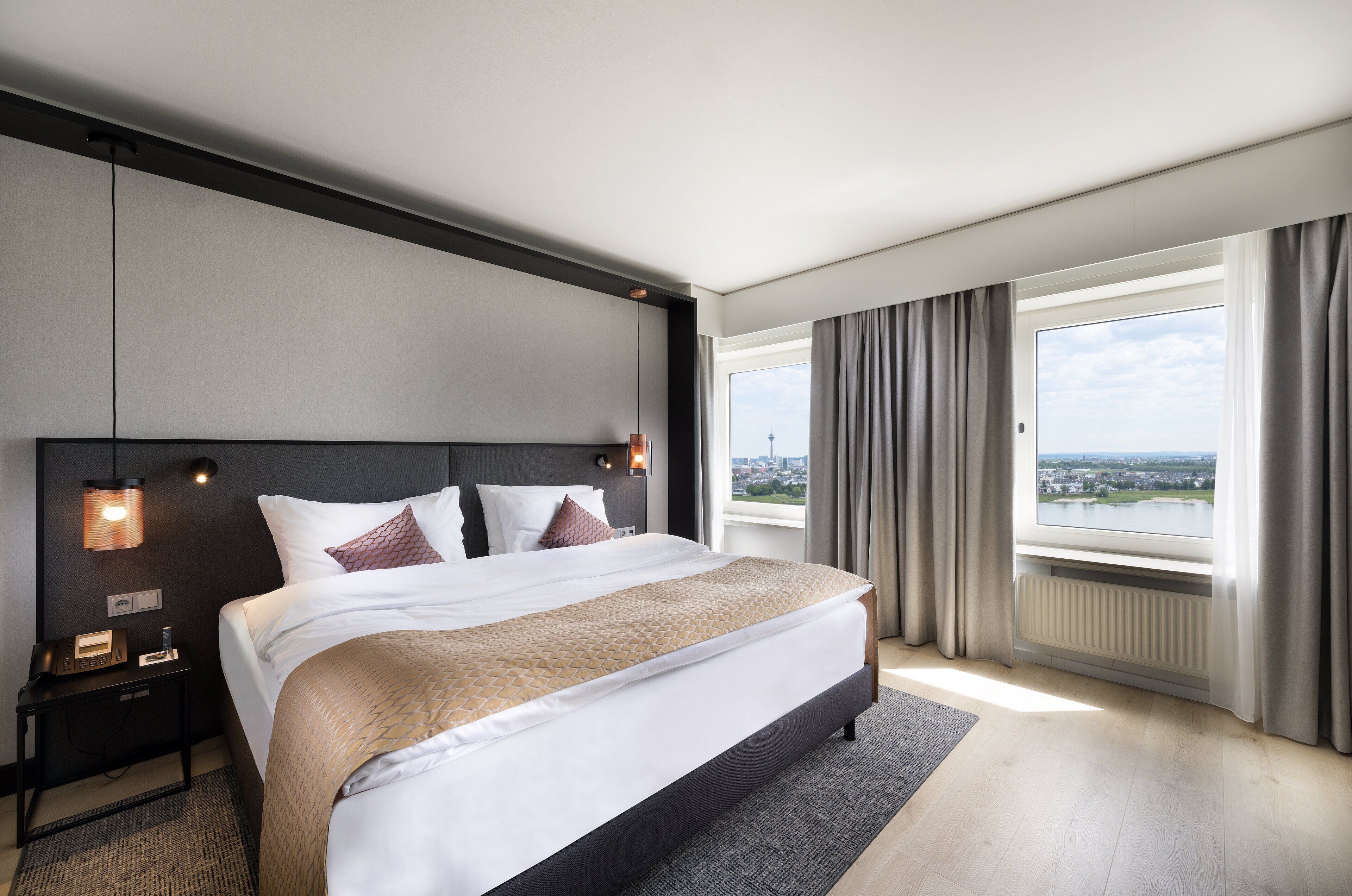 Una habitación de hotel moderna con una cama grande, una mesita de noche con lámpara, vistas de la ciudad a través de la ventana y un paisaje urbano visible desde el exterior.