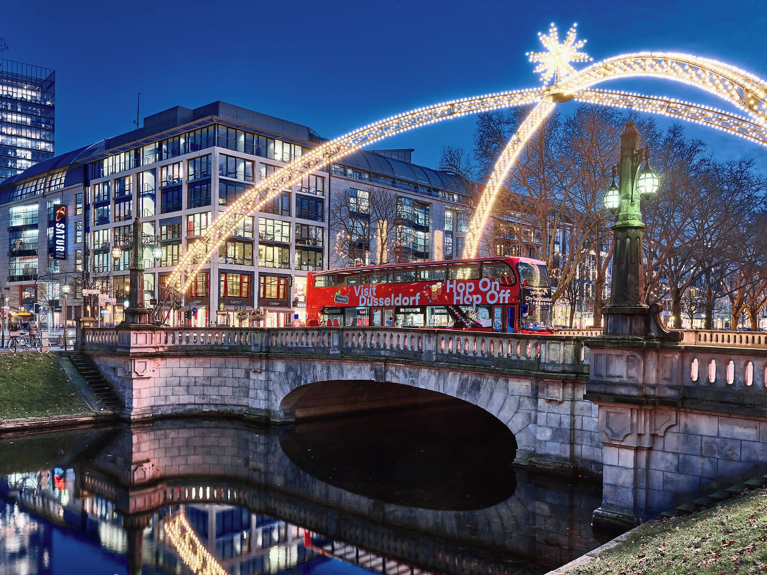 Ein roter, zweistöckiger Bus mit der Aufschrift 'Visit Düsseldorf Hop On Hop Off', eine Brücke und ein Spiegelteich.