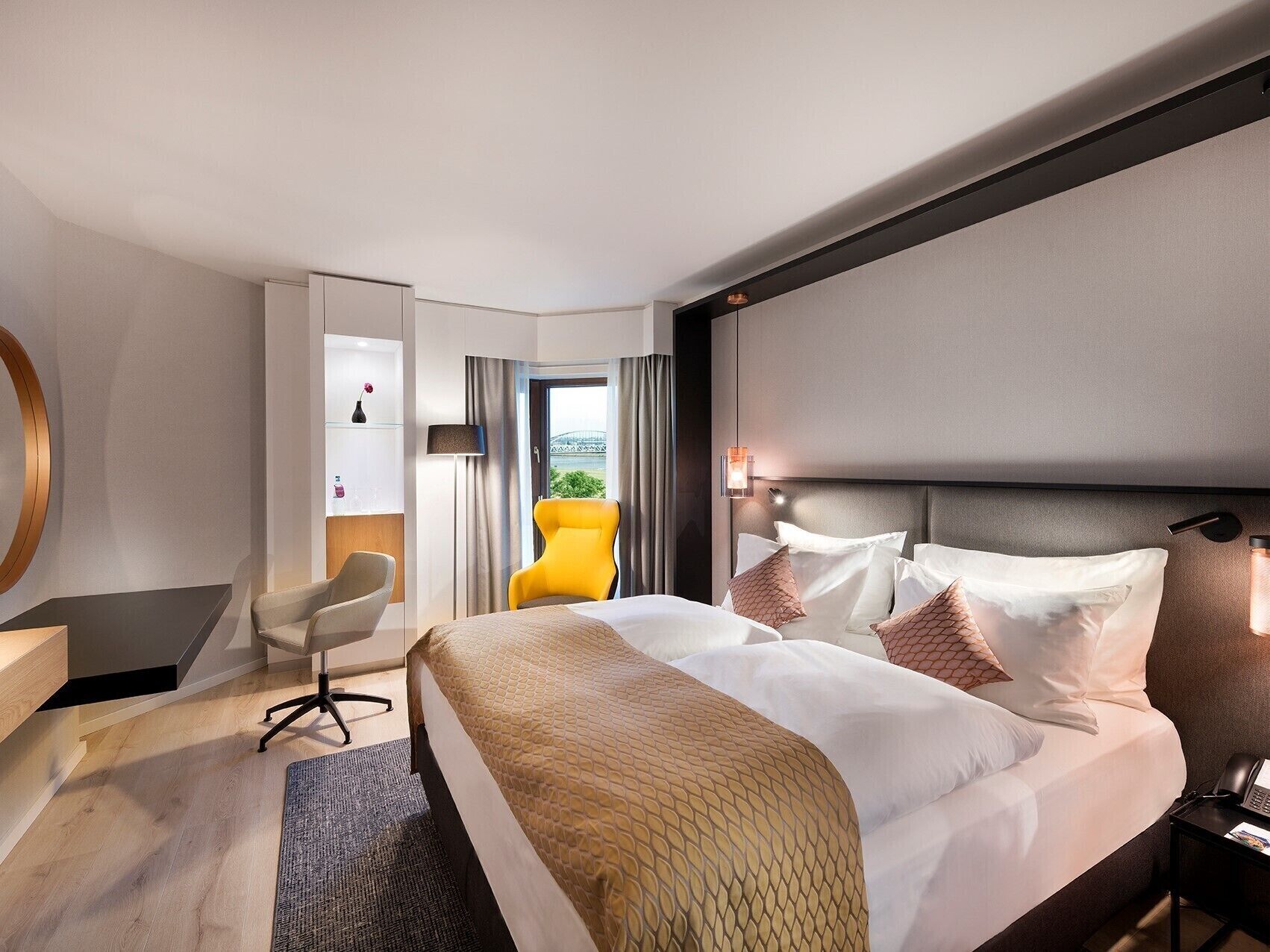 A modern hotel room with a large bed, a desk, a yellow chair, and a view of the outdoors.