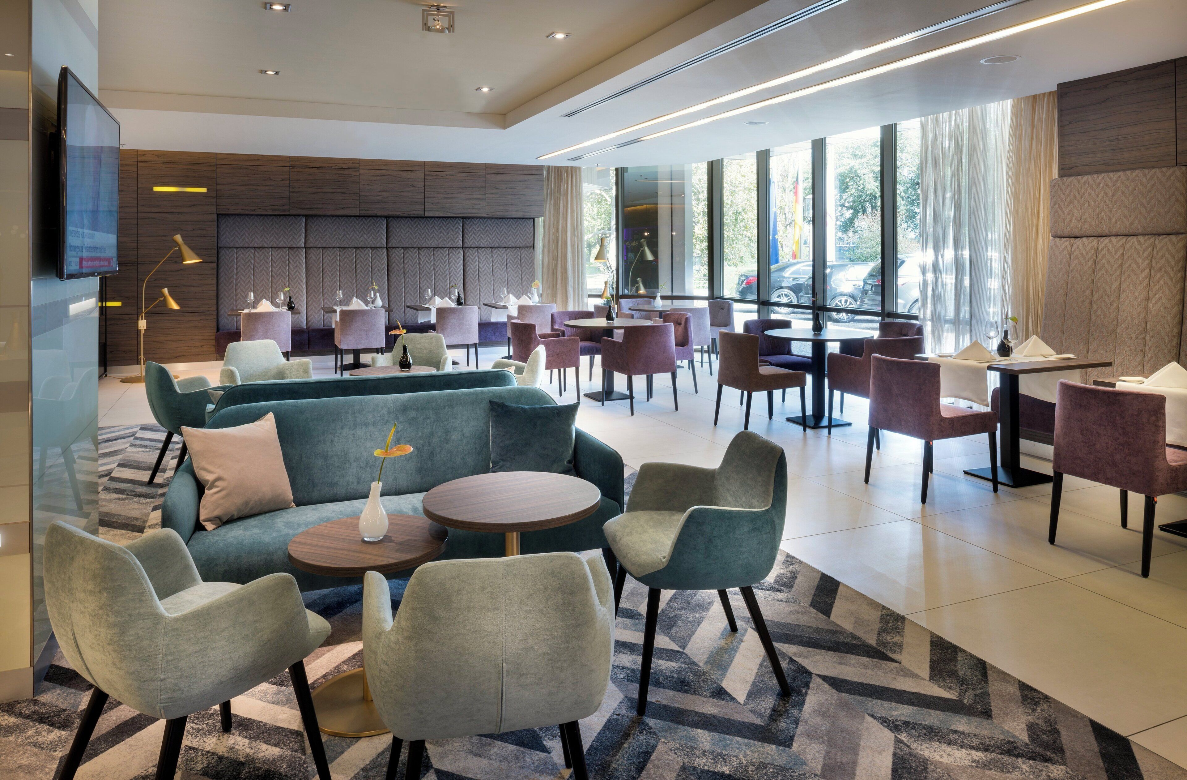 A modern lounge area with sofas, chairs, and tables, large windows with a view of the street, and a flat-screen TV mounted on the wall.
