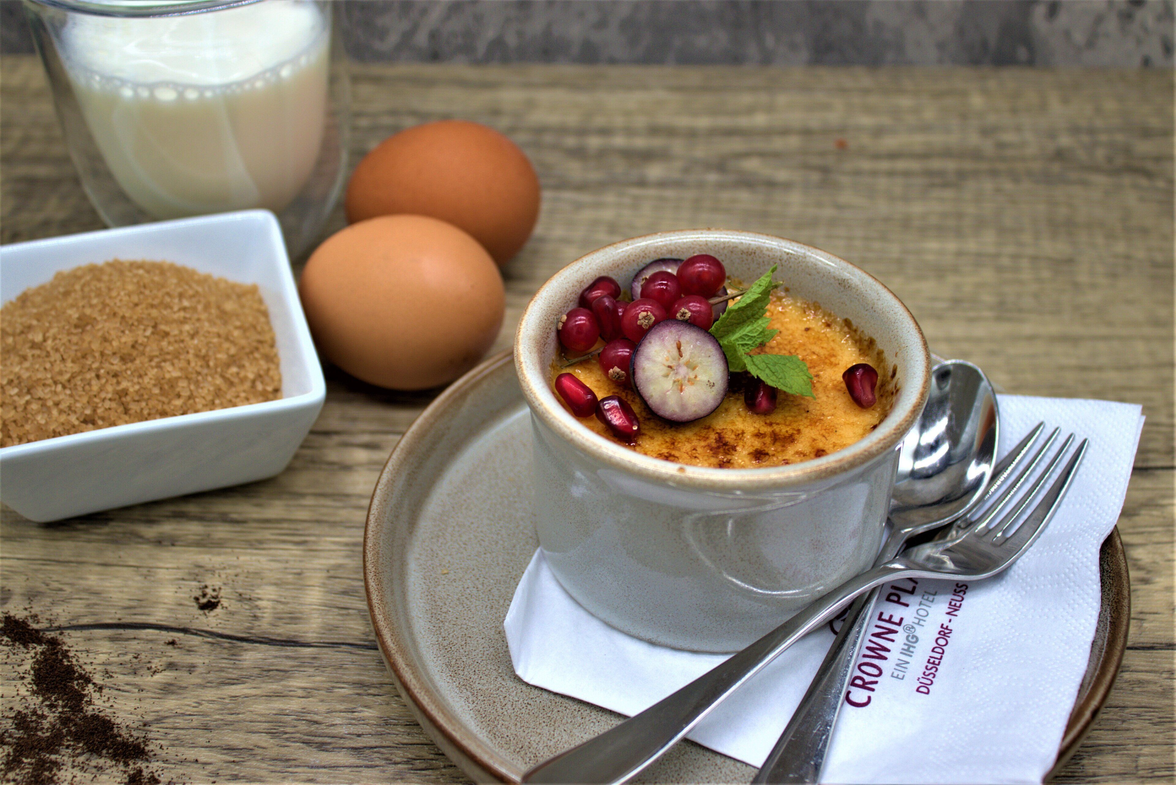 Ein Dessert im Glas mit Löffel und Gabel auf einem Untertaler, eine Schüssel mit braunem Zucker und zwei Eier auf einem Holztisch.