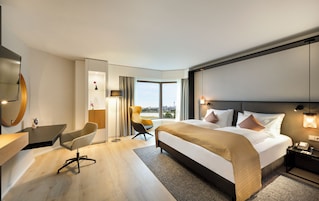 A modern hotel room with a large bed, a desk, a chair, and a view of the city.