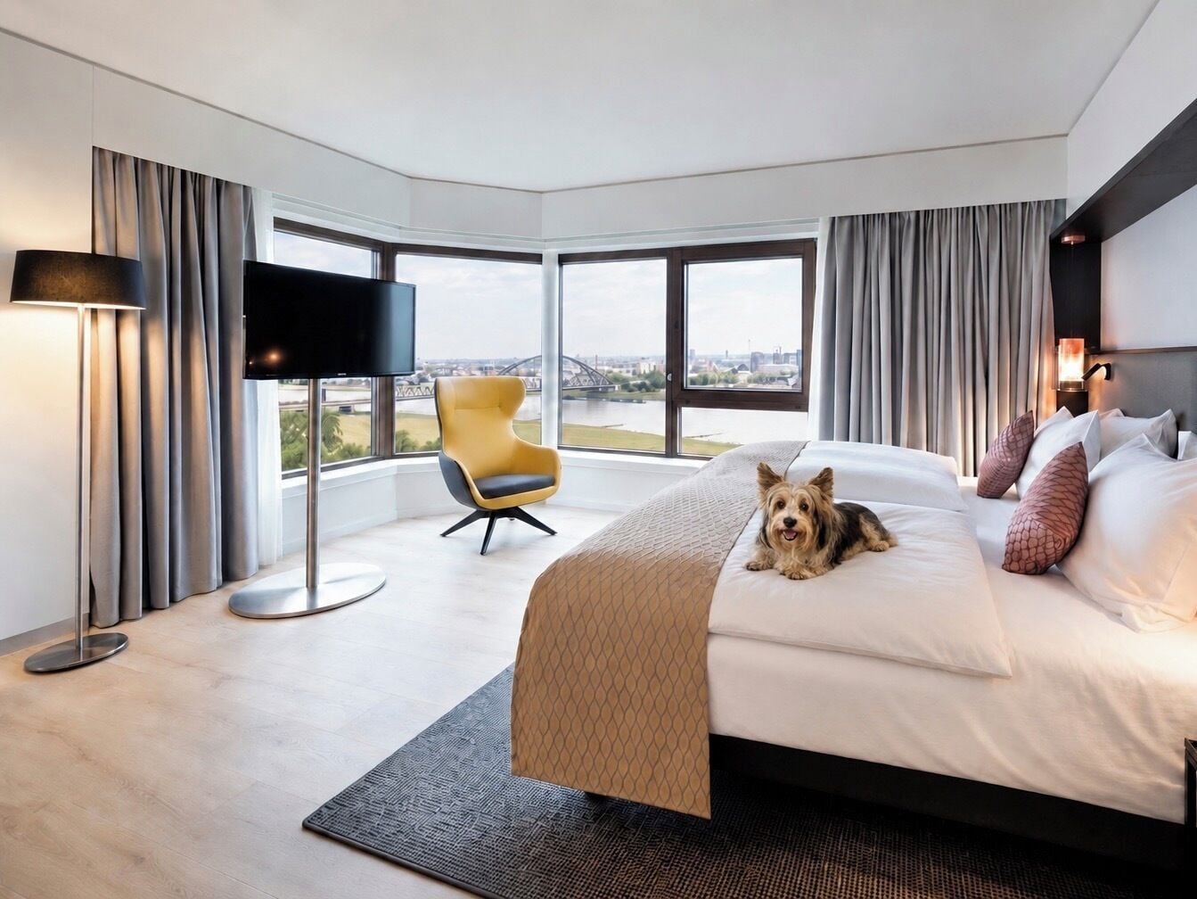 Una habitación de hotel moderna con una cama grande, un televisor de pantalla plana, un sillón amarillo y vistas a la ciudad.