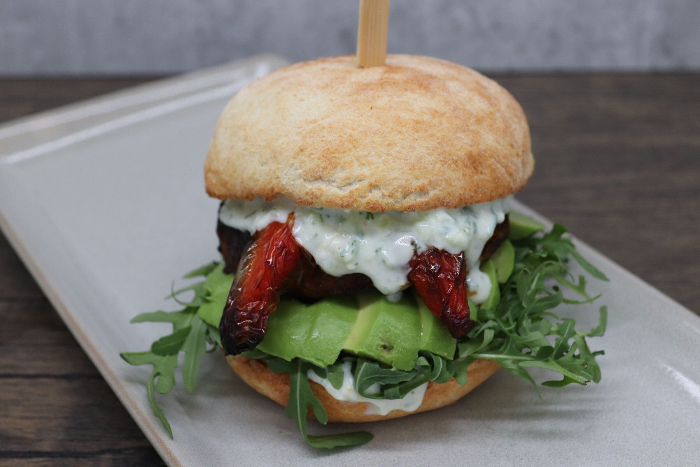 Un sandwich préparé avec un pain grillé, de l’avocat, des légumes verts et une sauce crémeuse, servi sur une assiette blanche.