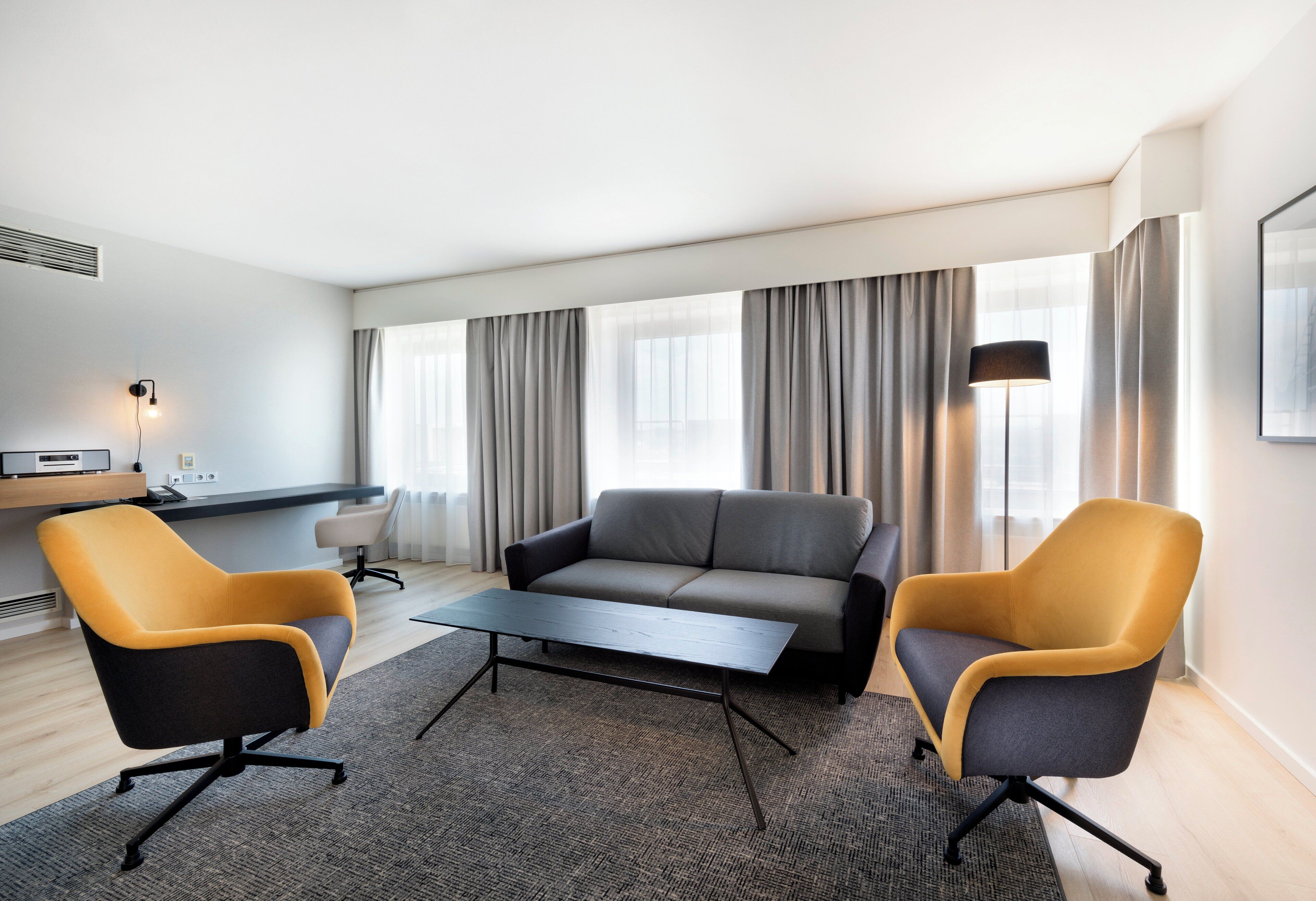 Una habitación de hotel moderna con un sofá gris, dos sillones amarillos, una pequeña mesa de centro y un escritorio con una lámpara.