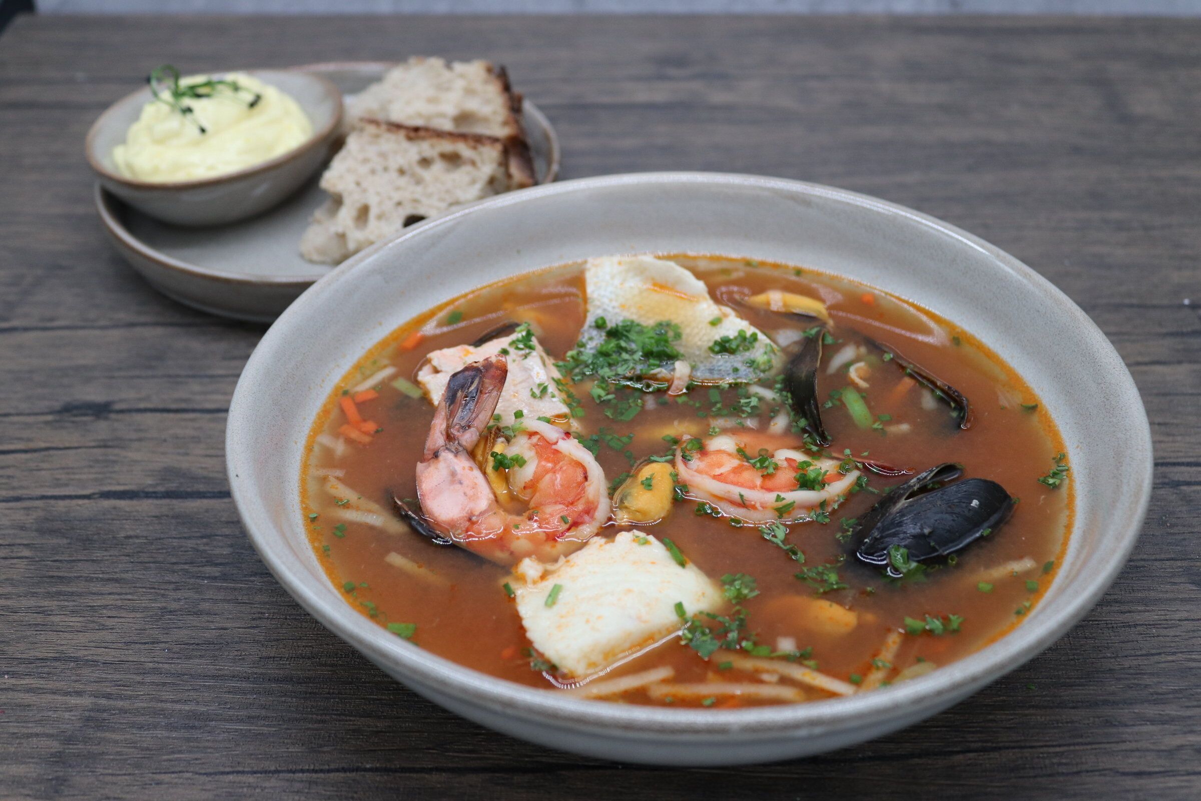 A bowl of seafood soup with various shellfish and vegetables, accompanied by a small bowl of sauce and slices of bread on a wooden table.