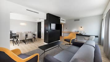 Ein modernes Wohnzimmer mit einer Couch, Sesseln, einem Essbereich mit Tisch und Stühlen sowie einer Küchenzeile mit Fernseher und einem kleinen Tisch.