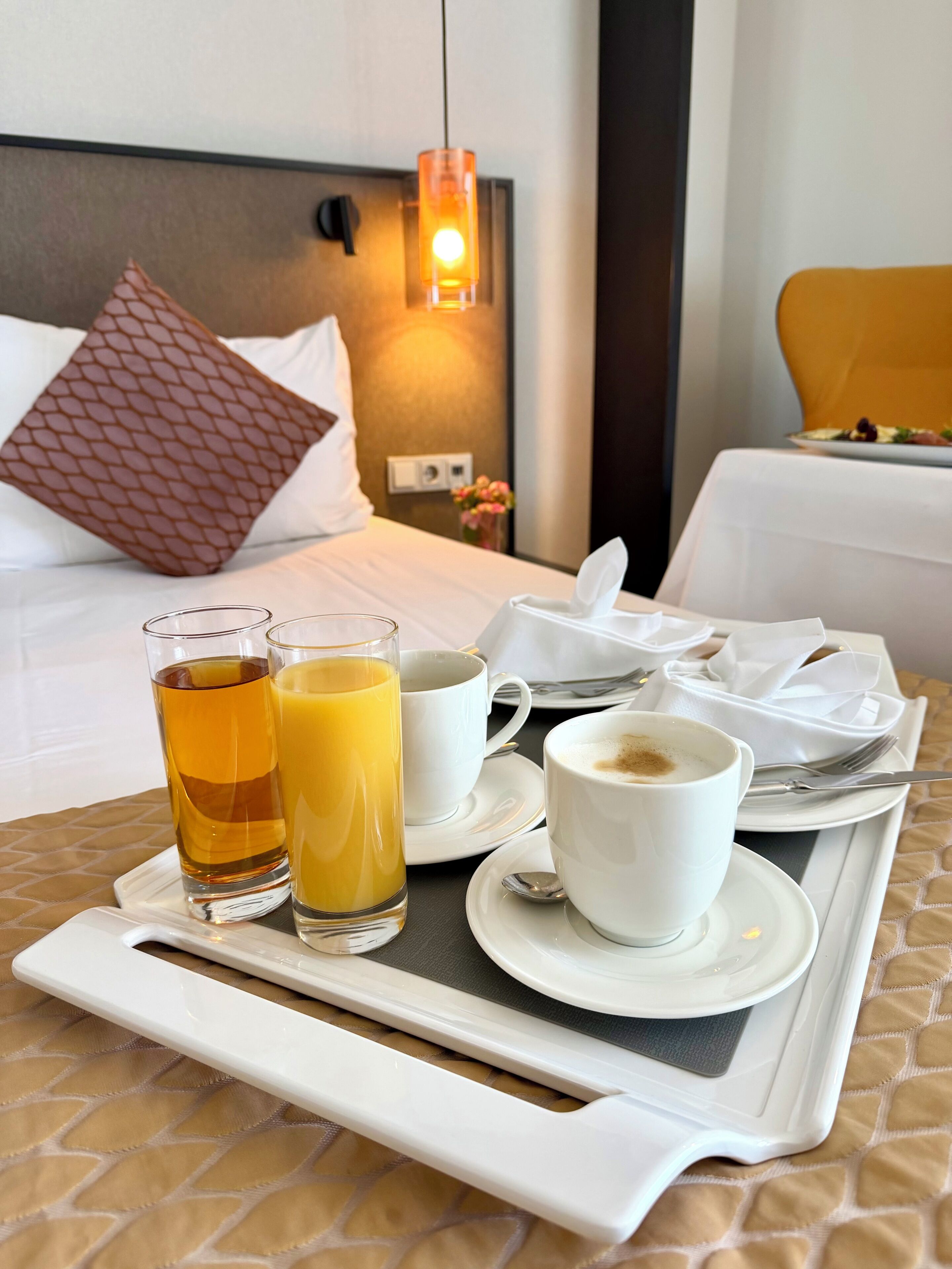 Una bandeja de desayuno ordenada con una taza de café, un vaso de té y un vaso de jugo de naranja sobre una cama con cabecera, almohadas y una mesita de noche con lámpara.