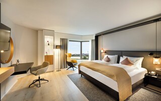 Une chambre d’hôtel moderne dotée d’un grand lit, d’un bureau, d’une chaise et offrant une vue sur la ville grâce à de grandes fenêtres.