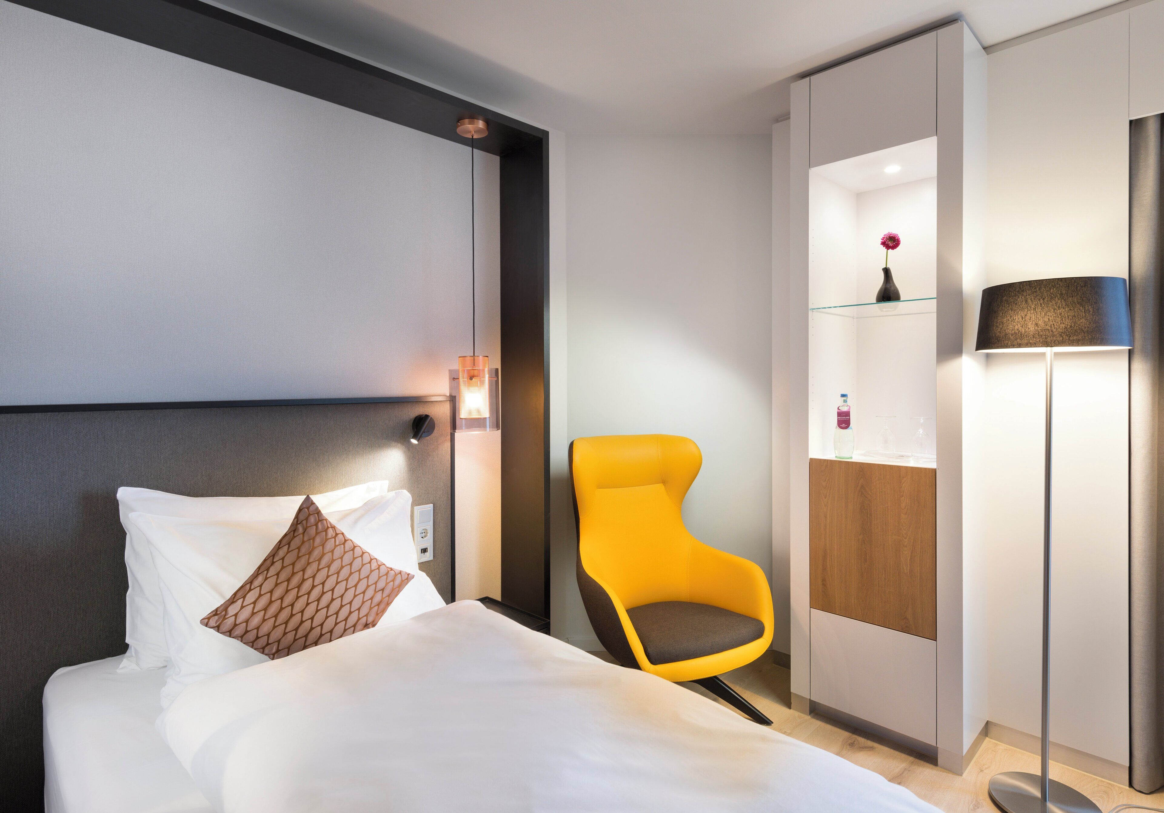 A hotel room with a bed, a yellow chair, a bedside table, a nightstand, and a wall-mounted lamp.