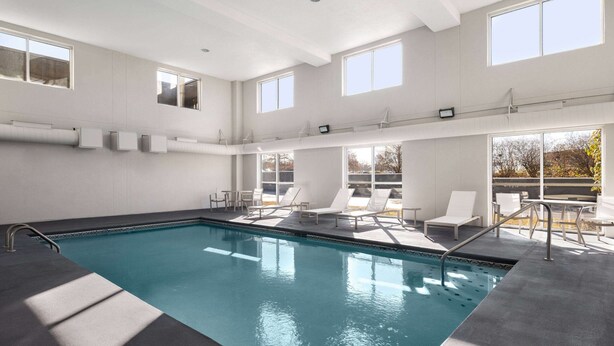 Ein moderner Poolbereich im Innenbereich mit freiem Blick durch große Fenster, ein Pool mit dunkler Kante und sonnendurchflutete Liegestühle.